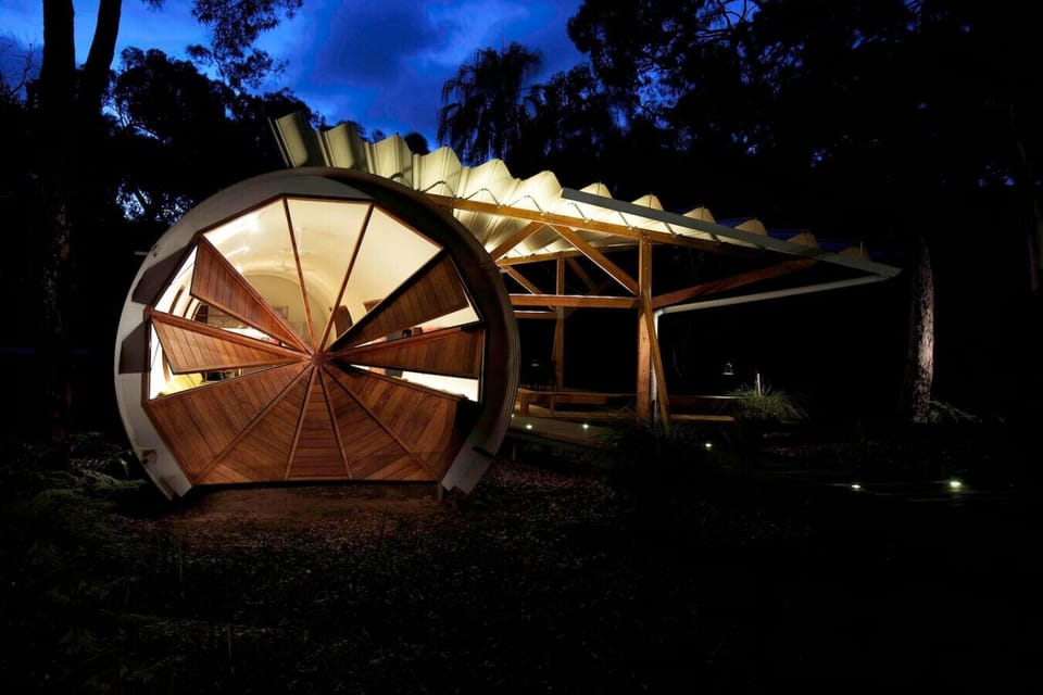 Contemporary pod exterior with cylindrical timber façade, canopy deck, and illuminated bushland setting.