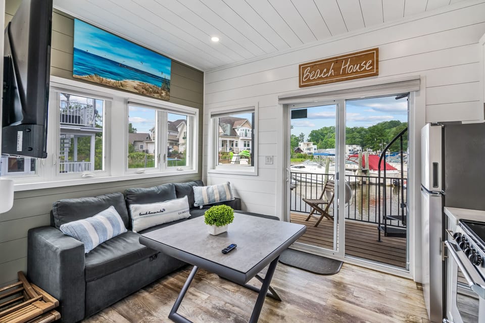 relax in the bright, waterfront living area at Shoreside Haven, featuring a comfy sofa, dining table, large windows overlooking the marina, and sliding doors that open to your private deck