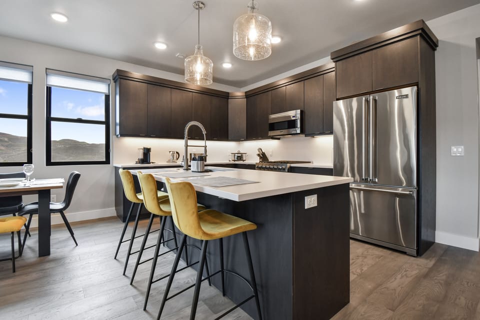 This modern kitchen boasts a spacious island with chic mustard-yellow barstools, ideal for casual meals or entertaining. Sleek cabinetry, stainless appliances, and airy windows elevate the space, blending style and function in an open-concept layout.