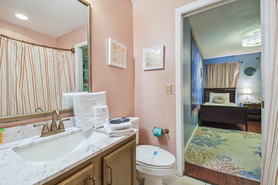 This bright bathroom boasts a sleek marble countertop and warm wooden cabinetry for a welcoming feel. Soft pastel walls enhance the tasteful decor, and an elegant shower curtain adds a charming touch for your comfort.
