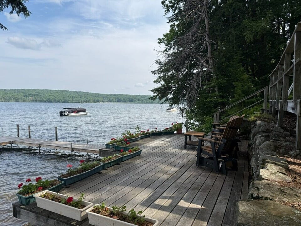Your private waterfront deck awaits, with Adirondack chairs perfectly positioned for morning coffee or evening relaxation overlooking the serene lake. Flowers, boat access, and wooded natural beauty surround you.