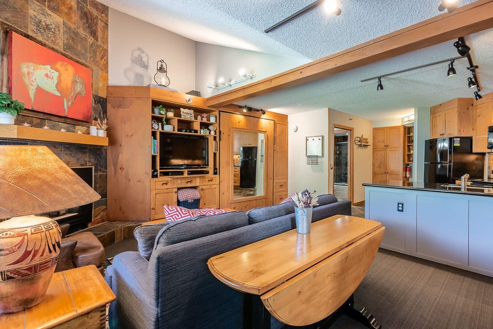 A cozy living room with wooden furniture, a fireplace, a wall-mounted TV, a painting of a cow, and track lighting. An adjoining kitchen features black appliances and an island with a countertop.