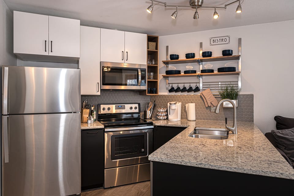 Modern kitchen with stainless steel appliances, white and black cabinetry, open shelving with black dishes, a granite countertop with a sink, and under-cabinet lighting.