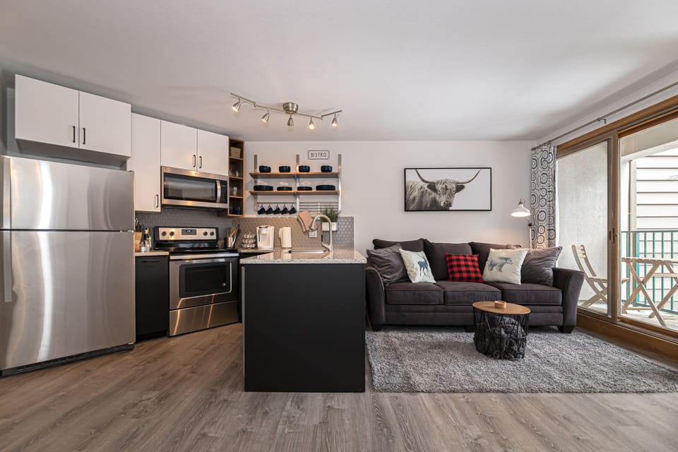 Modern studio apartment with an open kitchen featuring stainless steel appliances, a black countertop island, and white cabinets. Adjacent is a gray couch with plaid pillows, a wooden side table, and wall art.