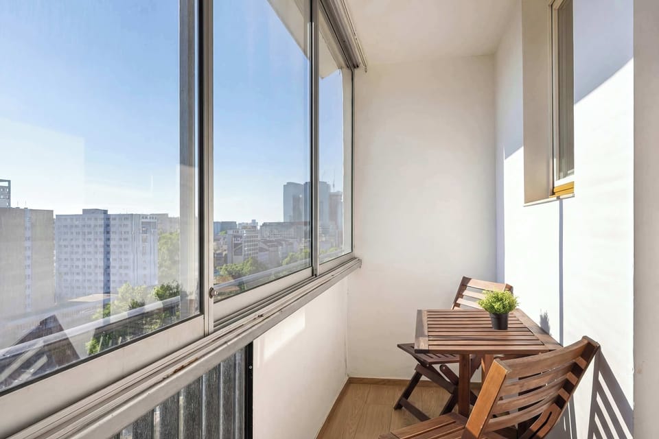 A balcony interior view with glass panels and a chair facing the city. Natural light fills the narrow outdoor space.