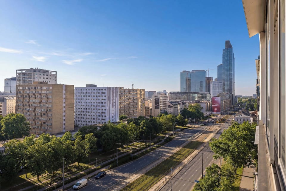 A city view from a higher floor showing rooftops, trees, and distant skyscrapers. The photo emphasizes urban density and greenery.