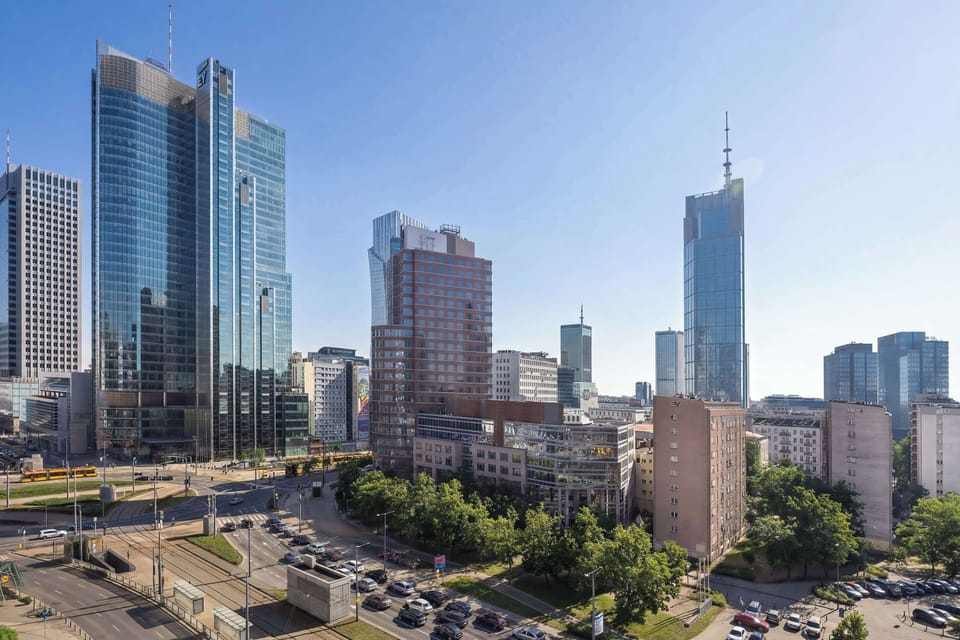 A city skyline view with modern high-rise buildings and busy streets below. The photo captures a bright daytime urban landscape.
