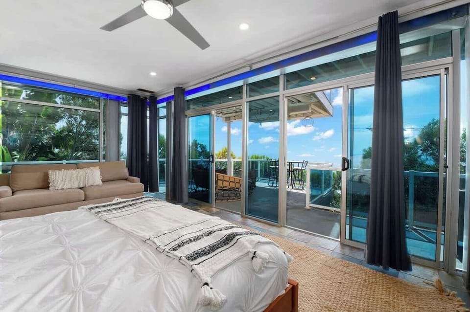 This room features a wraparound balcony with panoramic ocean views, thanks to the expansive glass doors and walls. A comfortable bed with plush pillows and the neutral rug create a serene atmosphere, perfect for unwinding after a day by the sea. 