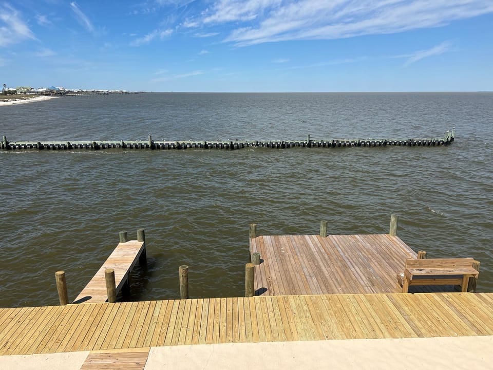 Waterfront deck with private dock stretches over calm bay waters beneath expansive blue skies.