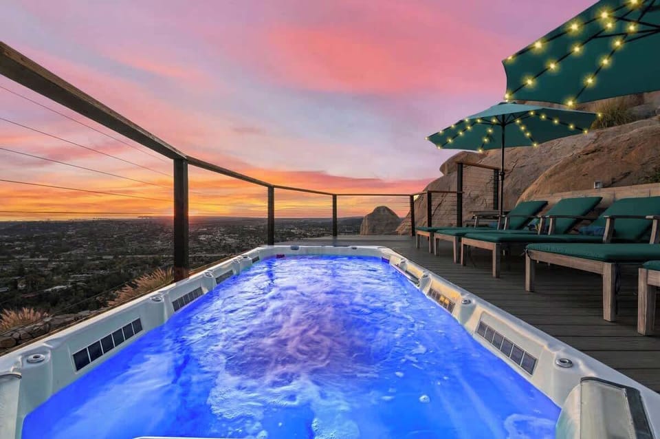 An inviting swim spa glows with clear water, perfectly positioned to capture a breathtaking sunset over the city. String lights hanging above the loungers create a magical ambiance, making this serene deck an ideal spot to take in the panoramic views