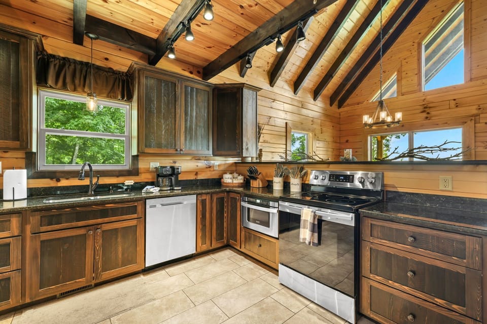 Open kitchen showcasing vaulted wood ceilings, exposed beams, stainless steel appliances, generous counter space, and large windows for natural light. Enjoy cooking in this Smoky Mountains cabin near Bryson City.