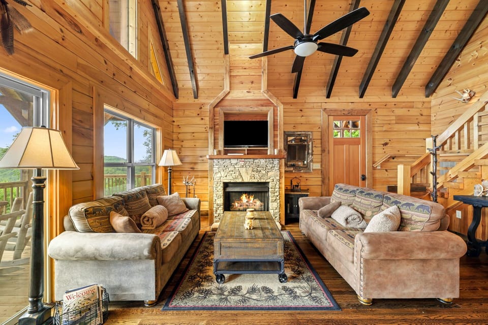 Spacious log cabin great room featuring vaulted wood ceilings, exposed beams, a stone fireplace, comfortable seating, and large windows overlooking the Smoky Mountains near Bryson City.