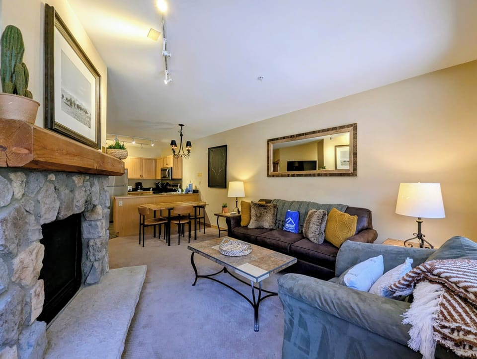 Picture yourself unwinding in this inviting living space, where a warm stone fireplace and plush seating create your perfect mountain retreat. Natural light fills the open layout, connecting your cozy lounge to a fully equipped kitchen and dining area.