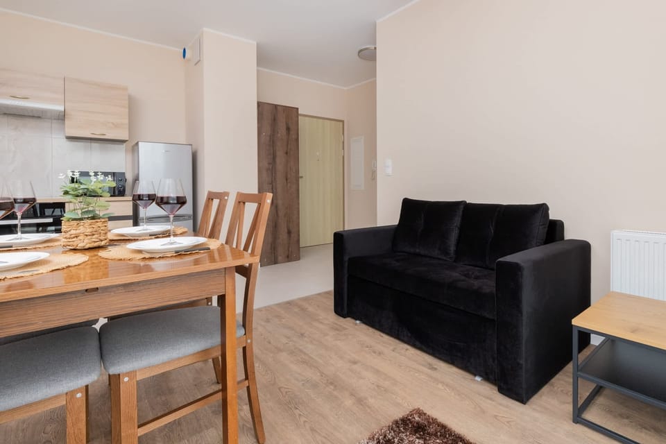 A bright and open living space with a cozy black sofa, a dining table for four, and large windows allowing natural light.
