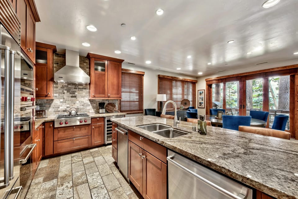 Granite island with double sink and leather barstools faces stainless steel appliances and brick tile backsplash.