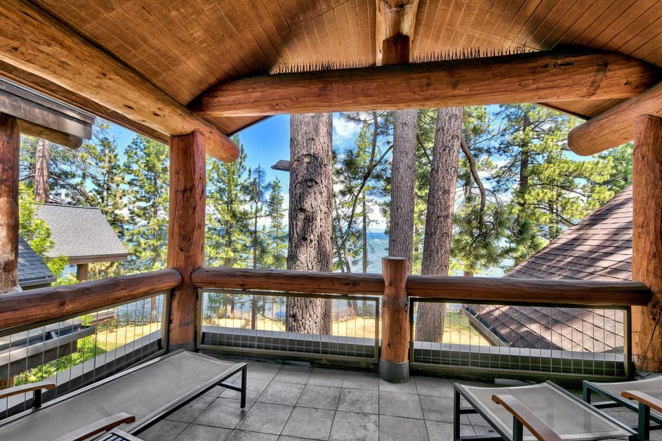 Covered balcony features large log beams, gray tiled floor, and mesh lounge chairs with lake and pine tree views.