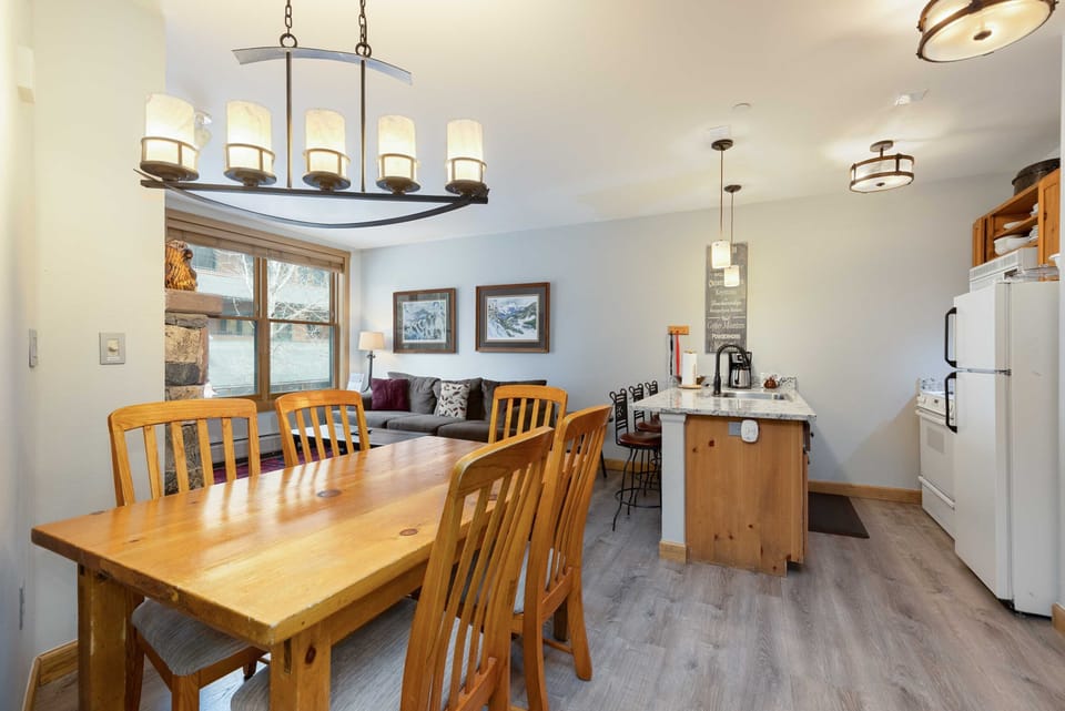 A cozy, modern kitchen and dining area with a wooden dining table, bench, pendant and ceiling lights, and a kitchen island. Decor includes framed art on the walls and a stone accent around the window.