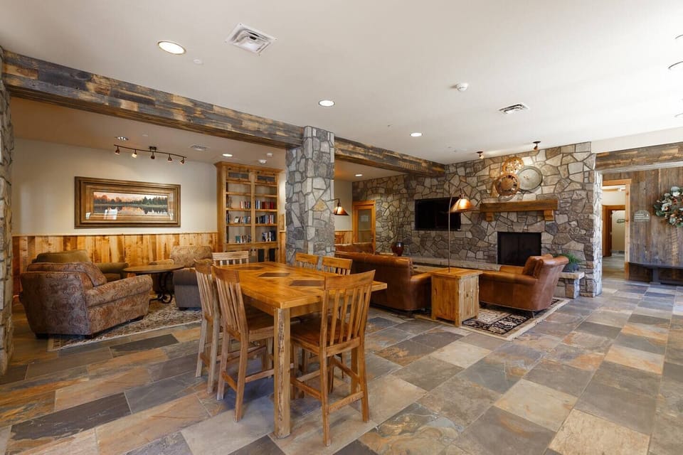 A spacious room with stone and wood walls, tile floor, a wooden dining table and chairs, several sofas, a large TV, a stone fireplace, and wooden bookcases filled with books.