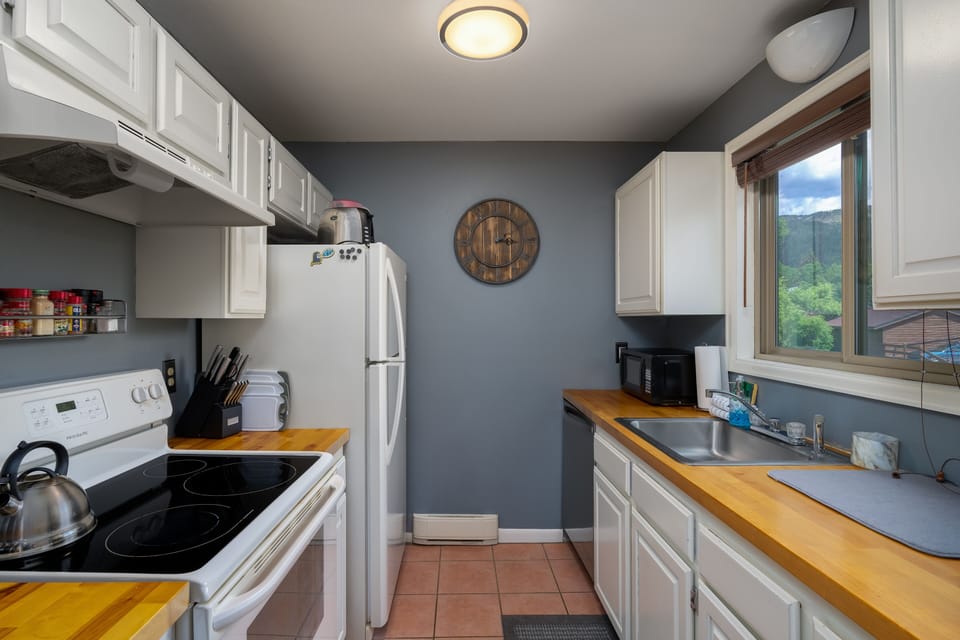 Butcher-block style countertops, induction top stove/oven combo, white and white refrigerator on the left side of the kitchen. Right side has stainless steel dishwasher, sink and longer section of countertop with white cabinets.