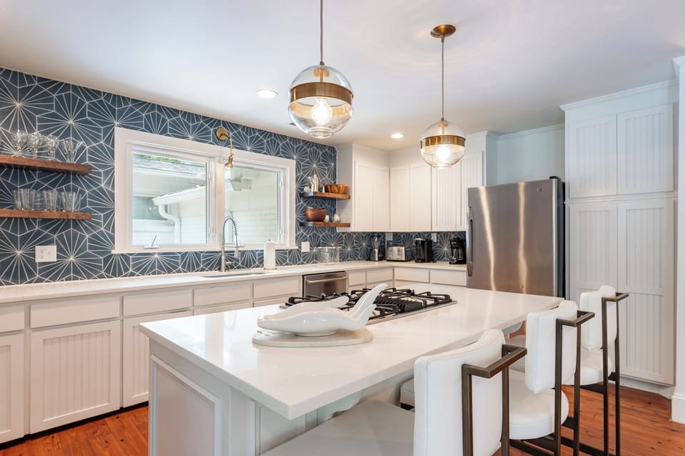 Open kitchen with white cabinets and countertops with wallpaper in dark blue. Glass pendant lights and stainless steel refrigerator.
