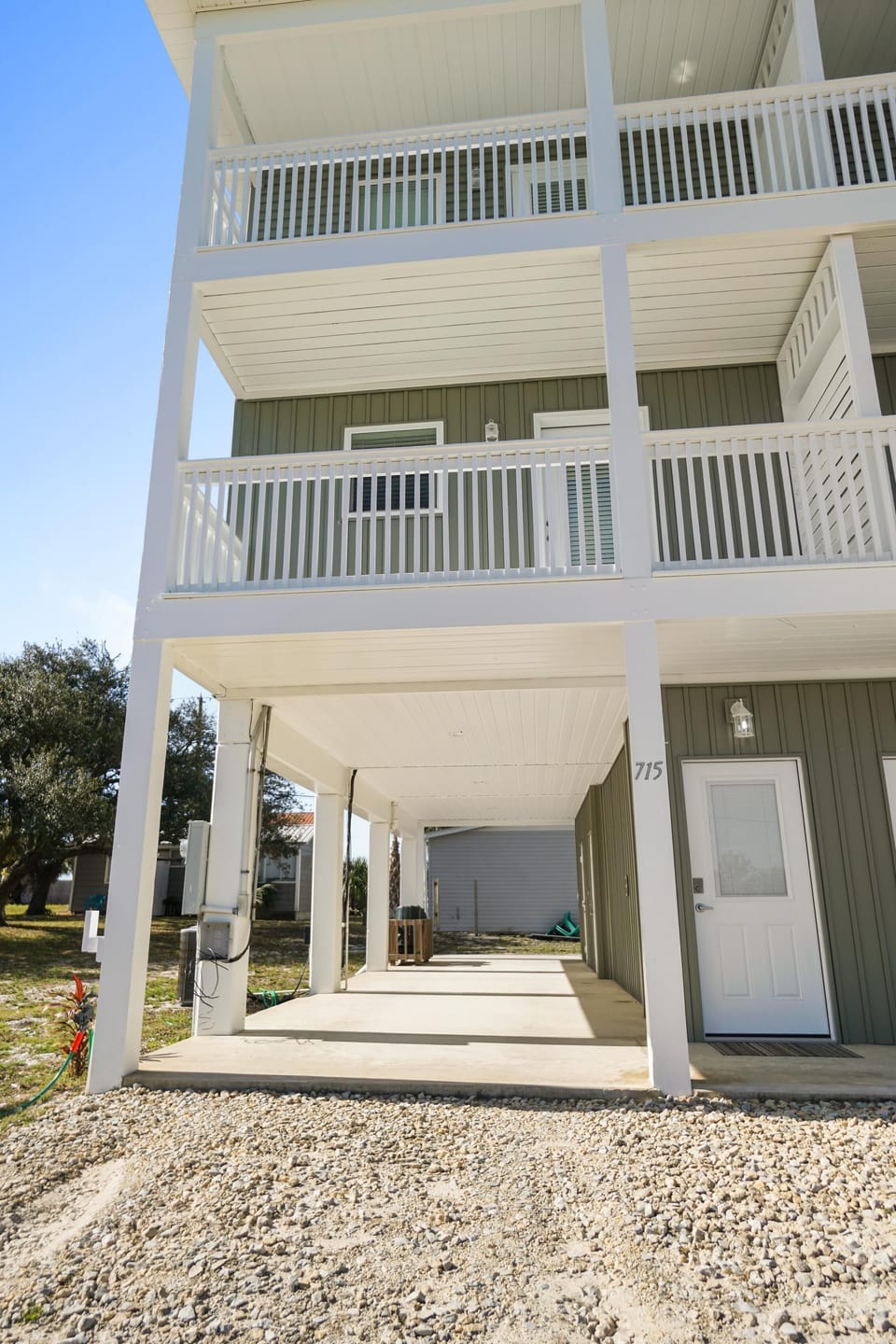Multi-story coastal building with white railings and covered ground-level entrance area, surrounded by natural landscaping.