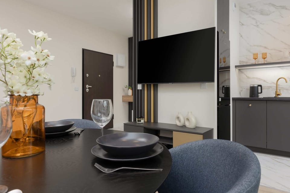 Close-up of a dining table with kitchenware and a decorative vase containing flowers. On the other ppa, a large flat-screen TV hangs on the wall.