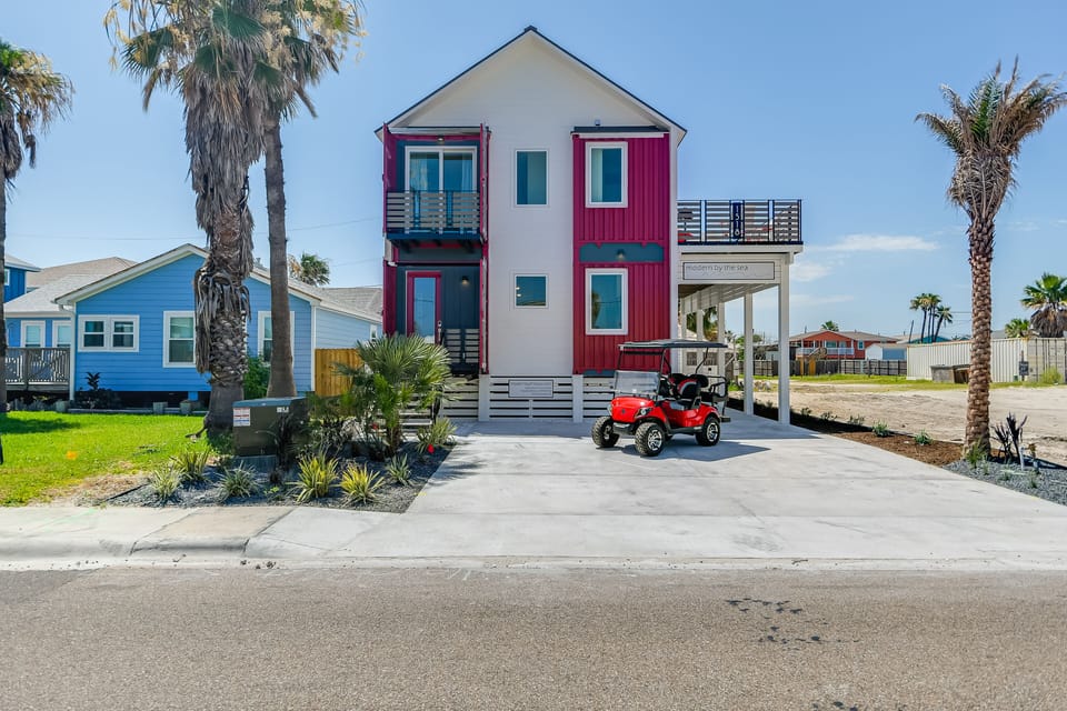 Modern beachside property featuring distinctive red and white architecture with covered parking and tropical landscaping in a coastal neighborhood setting.