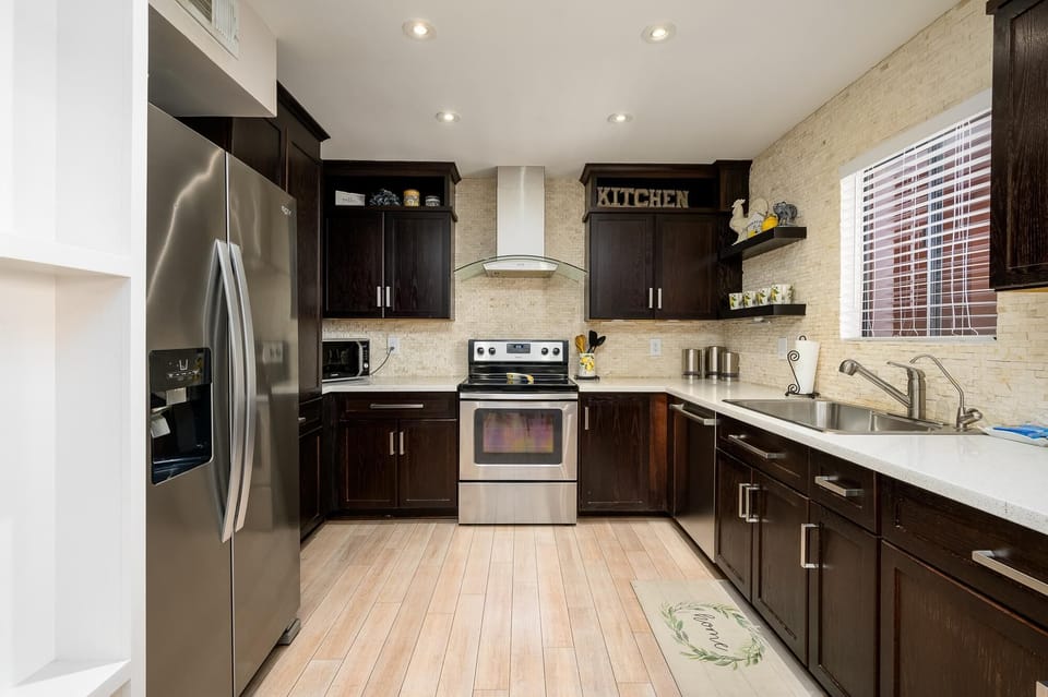 Recessed lighting brightens the kitchen with dark cabinetry, stainless steel sink, and light wood flooring.