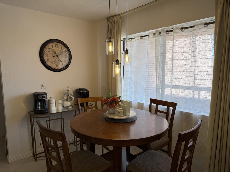 Round wooden dining table with four chairs beneath pendant lights and sheer curtains on the window.