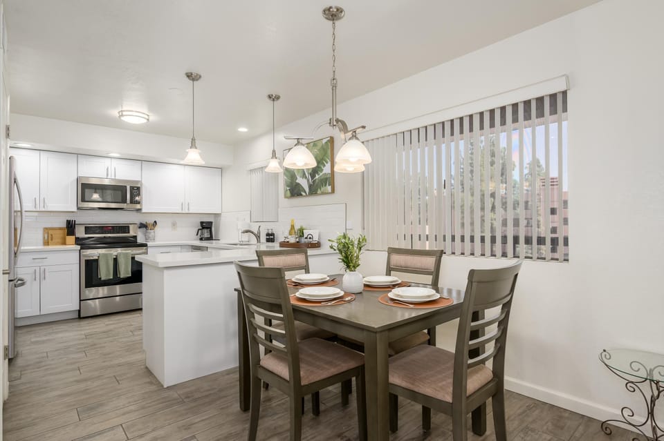 Square wood dining table set with four beige-cushioned chairs under pendant lights by large vertical window blinds.