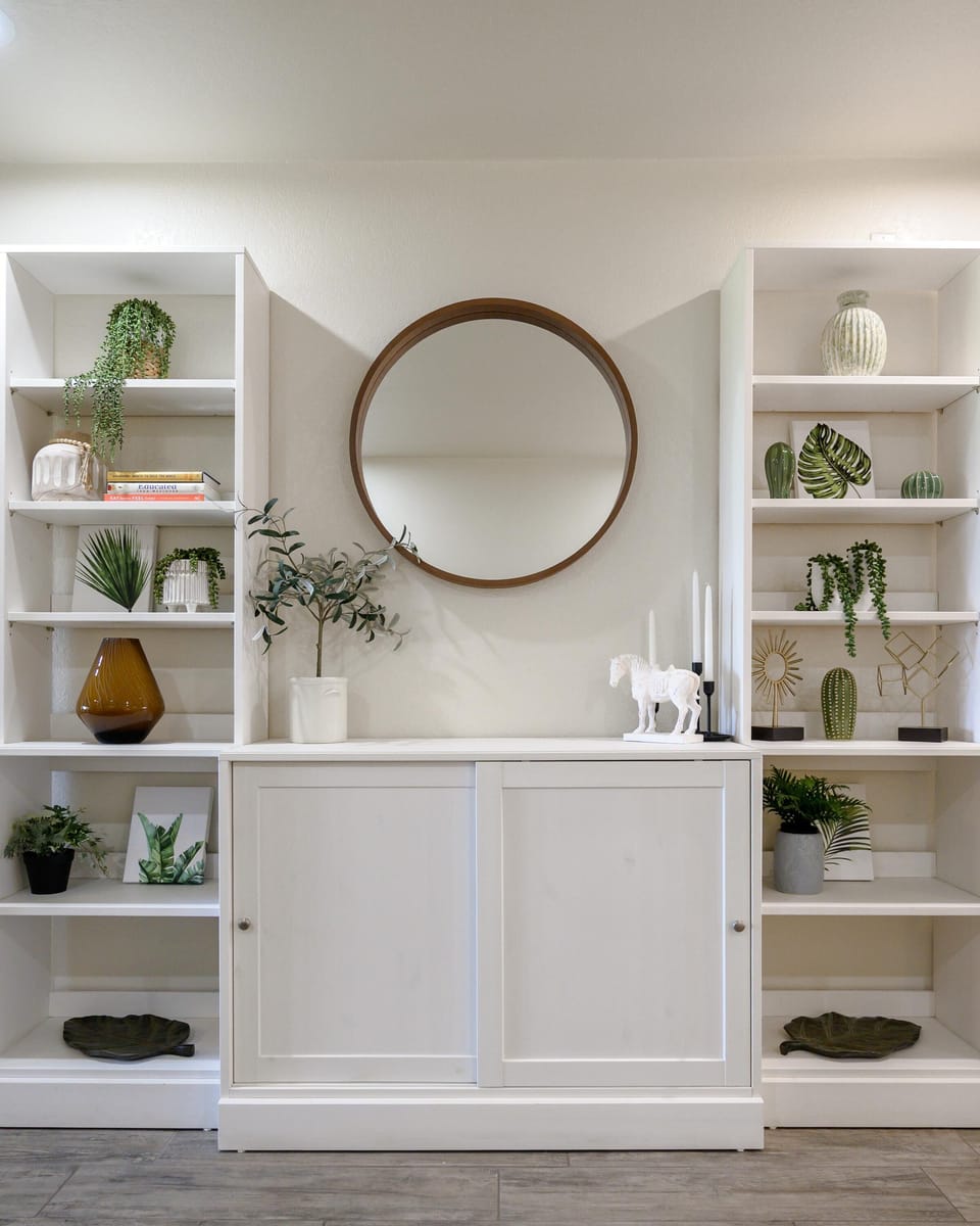 Built-in white shelving holds green plants, decorative vases, and framed art beneath a round wood-framed mirror.