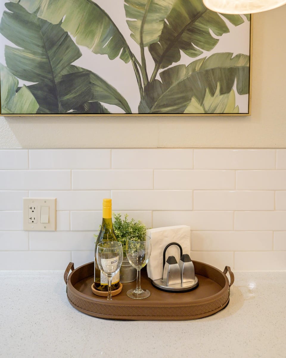 Kitchen countertop with white subway tile backsplash, a framed leaf print, and a serving tray with wine and glasses.