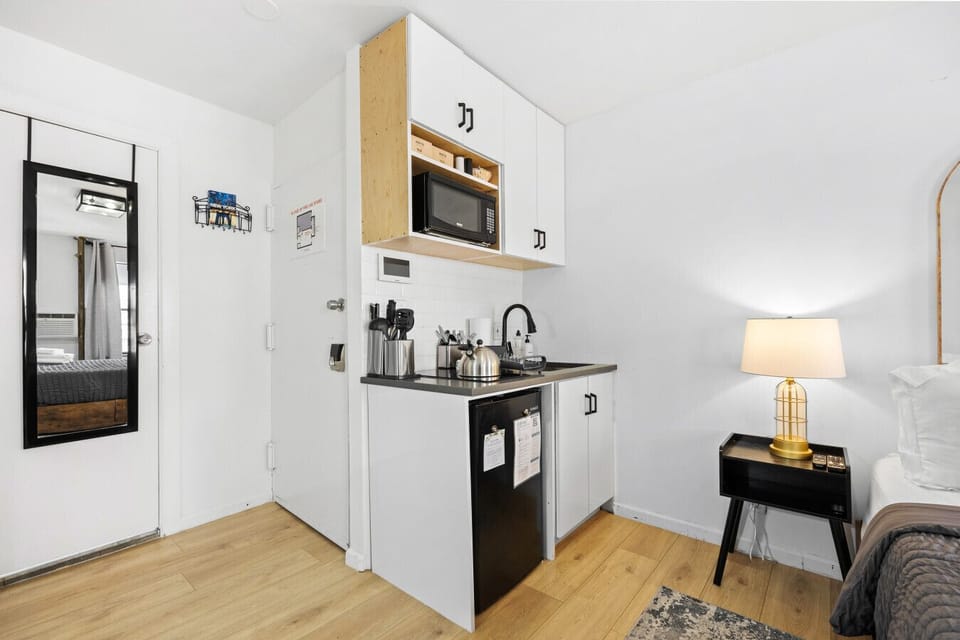 Compact kitchenette with white cabinets, sink, microwave, mini fridge, kettle, and utensils. Mirror near the door.