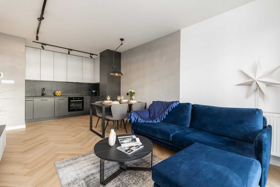 A modern, open-plan living room with a blue sofa, a dining table with chairs, and a large window letting in natural light.