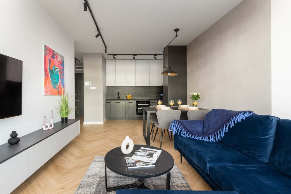 A modern, open-plan living room with a blue sofa, a dining table with chairs, and a large window letting in natural light.