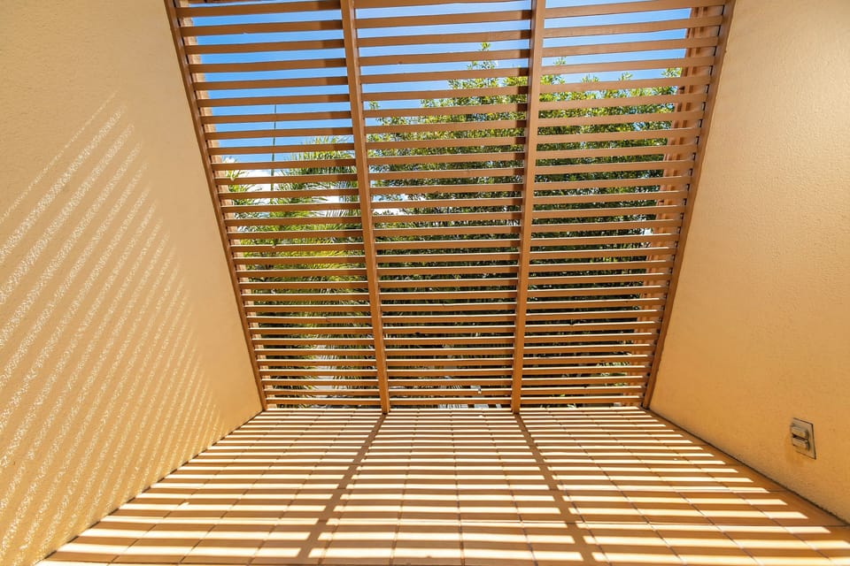 Sunlight filters through slatted wooden screens overhead, casting dramatic shadows across the tiled floor and warm walls of your private outdoor space.