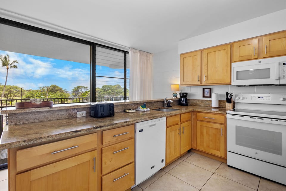 Your fully-equipped kitchen awaits with modern appliances, warm cabinetry, granite countertops, and stunning tropical views through expansive windows—perfect for preparing meals while soaking in the scenery.