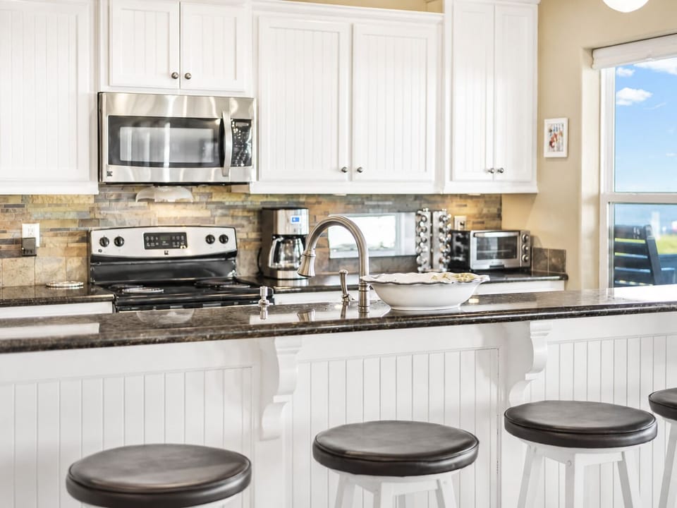 Modern coastal kitchen featuring granite countertops, white cabinets, stainless steel appliances, deep sink, and a breakfast bar with stools.