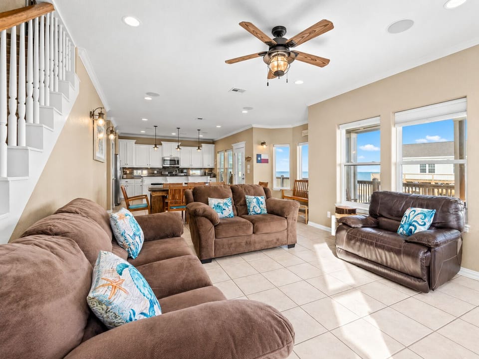 Sunlit open living room with multiple sofas, ceiling fans, large windows, staircase access, and plenty of comfortable seating for guests.