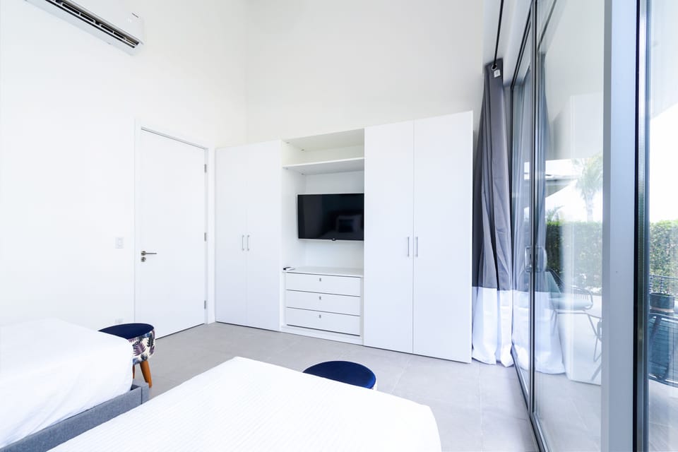 Your bright, airy bedroom features clean white wardrobes with built-in TV, modern air conditioning, and floor-to-ceiling glass doors opening to your private balcony. Stylish seating and ample storage complete your peaceful retreat.