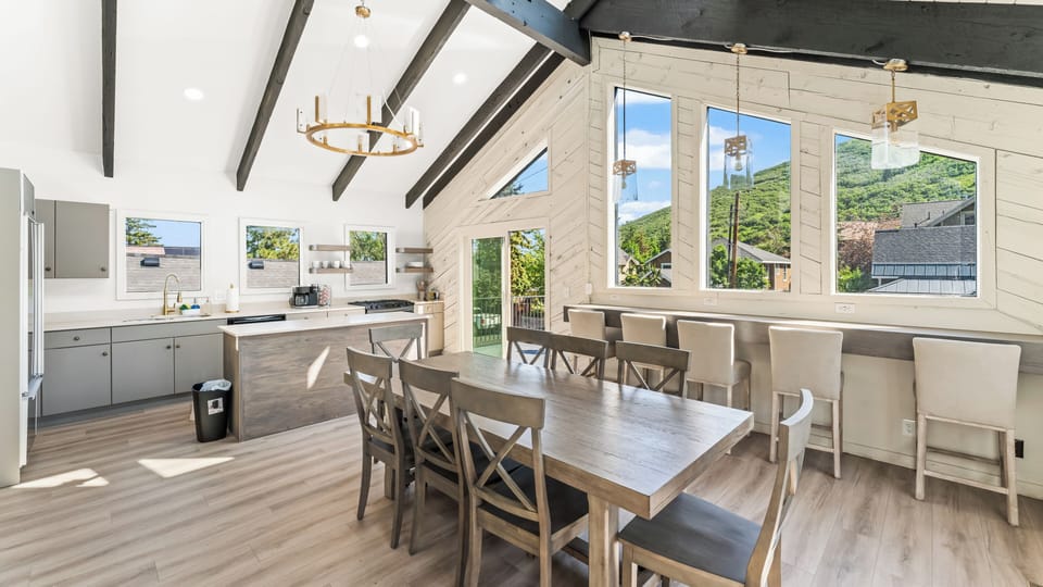 - Open kitchen and dining with vaulted ceilings and beams  
- Long wooden dining table plus breakfast bar seating  
- Expansive windows highlight mountain and forest views