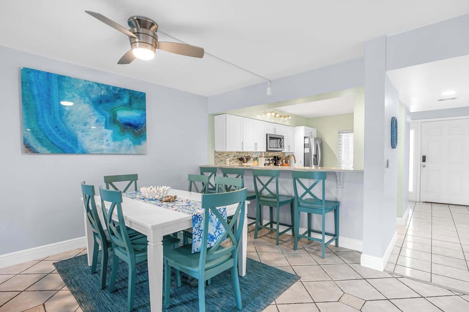 Dining area with teal chairs and a modern ceiling fan adjacent to a kitchen with white cabinetry.