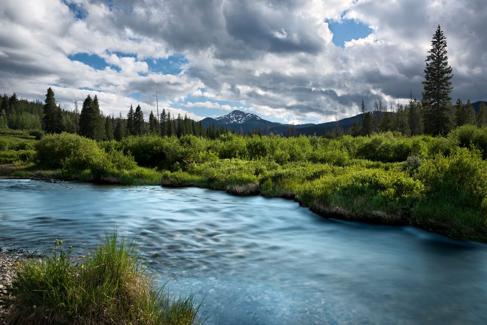 A pristine river winds through a lush valley framed by evergreen forests and dramatic mountain peaks, capturing the serene natural beauty surrounding the property.