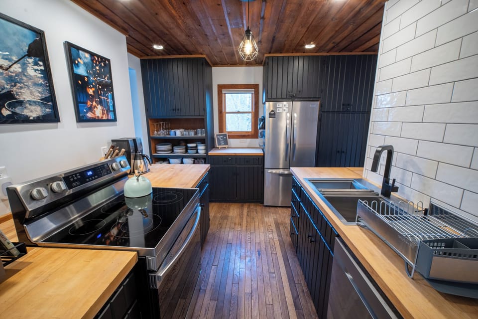 Modern galley kitchen with dark cabinets, butcher block countertops, stainless steel appliances, and a vaulted wood ceiling.