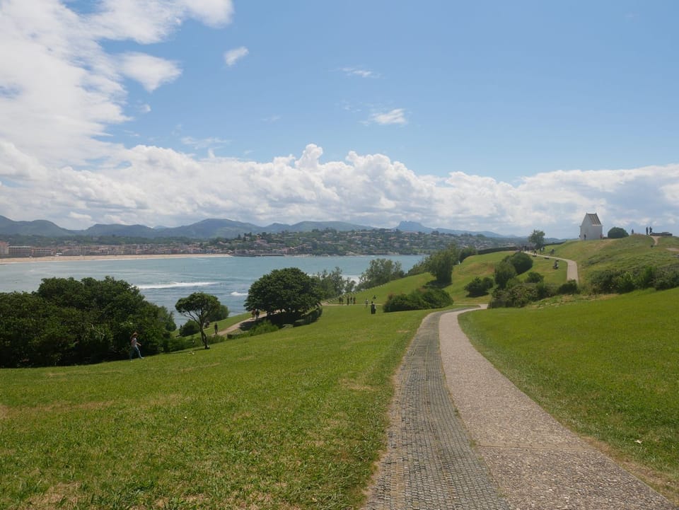 A winding path leads across the grassy slopes of Sainte-Barbe headland, with sweeping views over Saint-Jean-de-Luz bay and the Pyrenees beyond.
