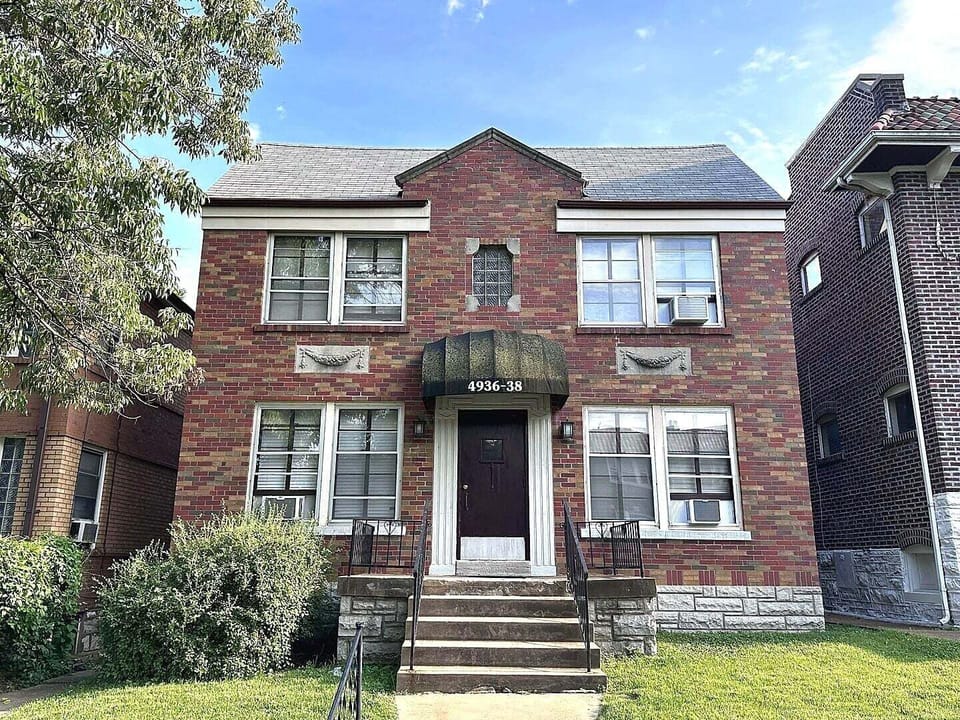 The daytime exterior shows the two-story brick building marked 4936-38, its central entrance and stairs, front lawn, mature tree, and neighboring residences.