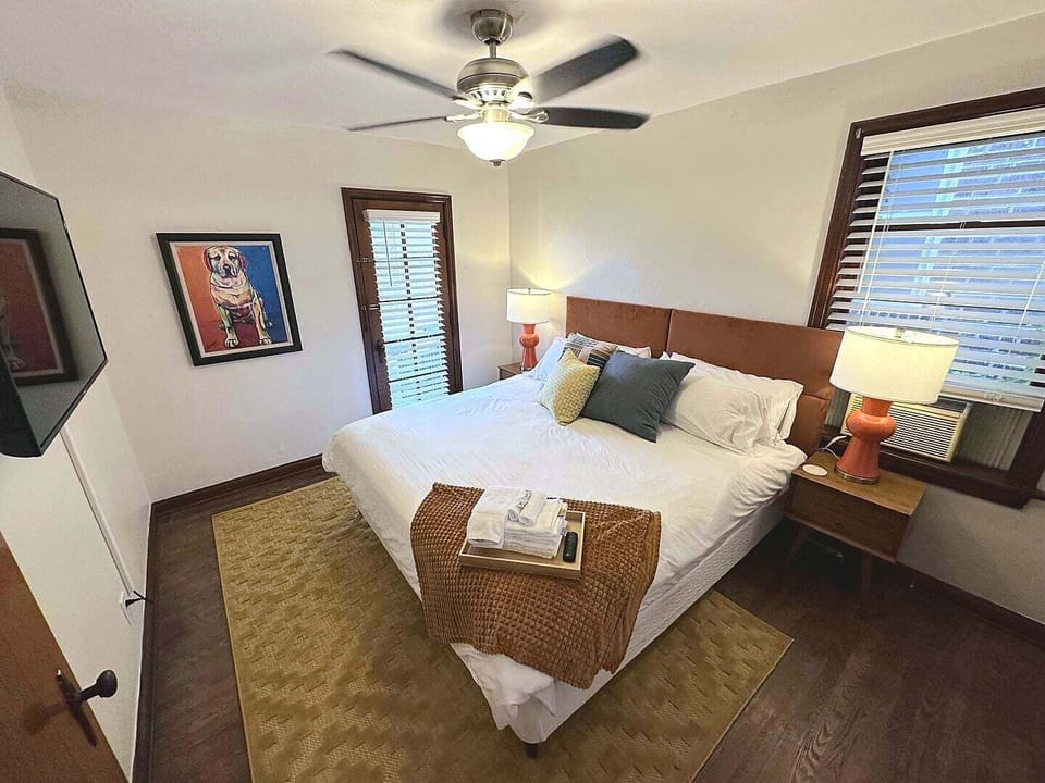 The primary bedroom includes a white-dressed bed, upholstered brown headboard, wall-mounted TV, ceiling fan, window with AC, nightstands, lamps, and a windowed door.