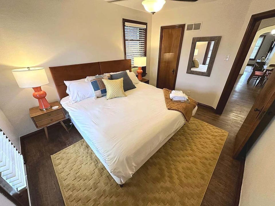 This primary-bedroom angle shows the white-dressed bed, brown upholstered headboard, nightstands, lamps, ceiling fan, closet, wall mirror, and doorway toward the adjoining space.