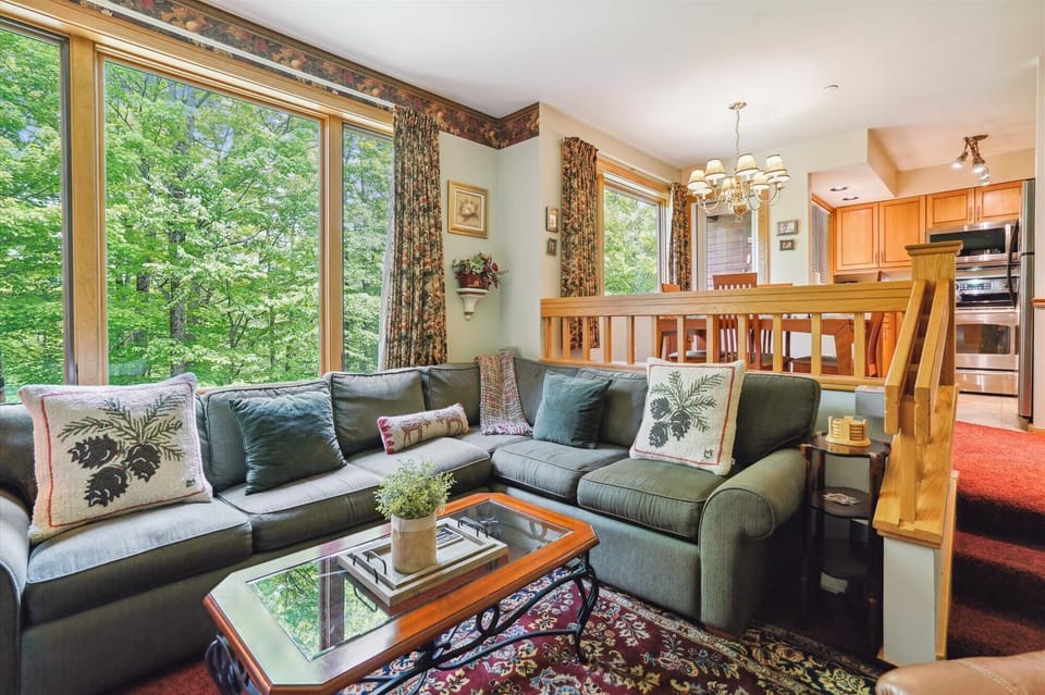 Inviting living room filled with natural light and surrounded by wooded views. Features a spacious sectional and open layout connecting to the dining and kitchen areas.