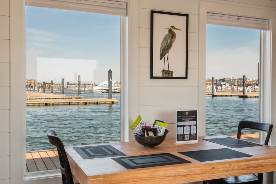 Four-seat dining table set against picture windows framing Lighthouse Point Marina, with heron artwork and natural light pouring in across the Patapsco River.